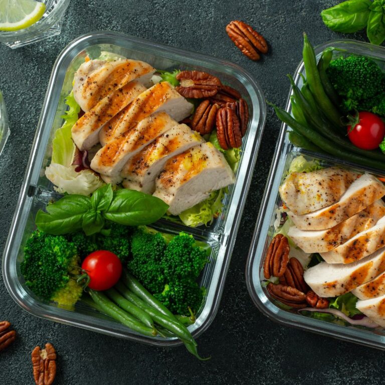 Two meal prep containers filled with grilled chicken, green beans, broccoli, pecans, and fresh greens.