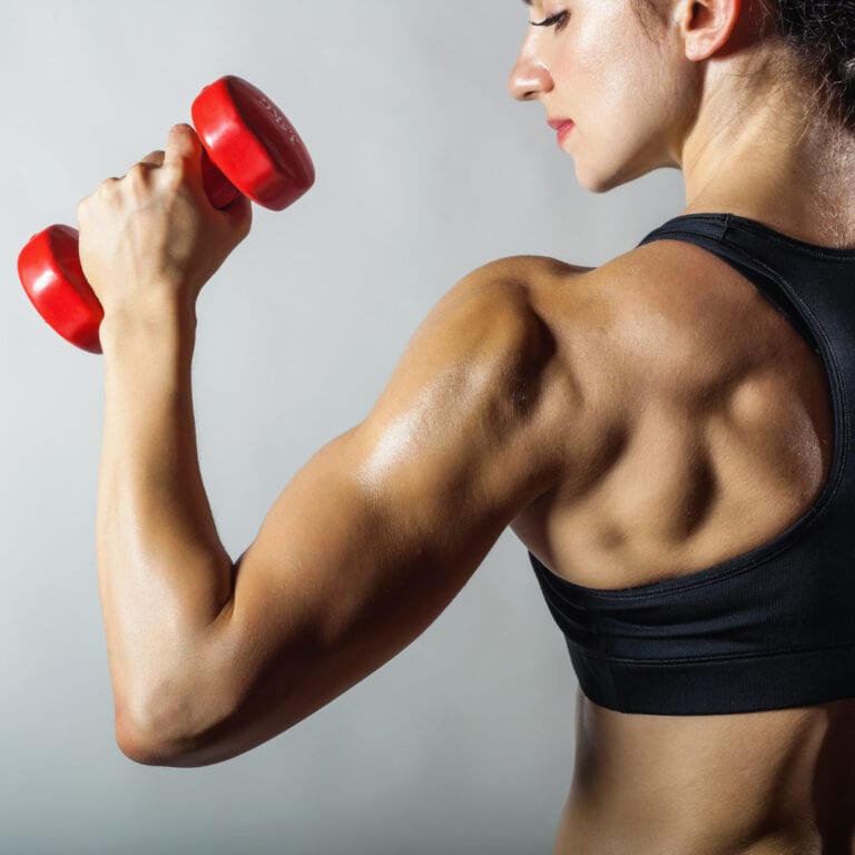 A fit woman in a black sports bra performing a bicep curl with a red dumbbell, showcasing her toned back and arm muscles.