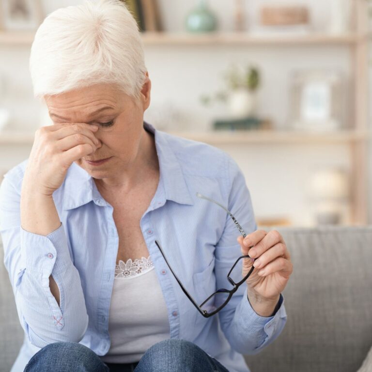 An older woman sitting on a couch, holding her glasses and rubbing her forehead in discomfort.