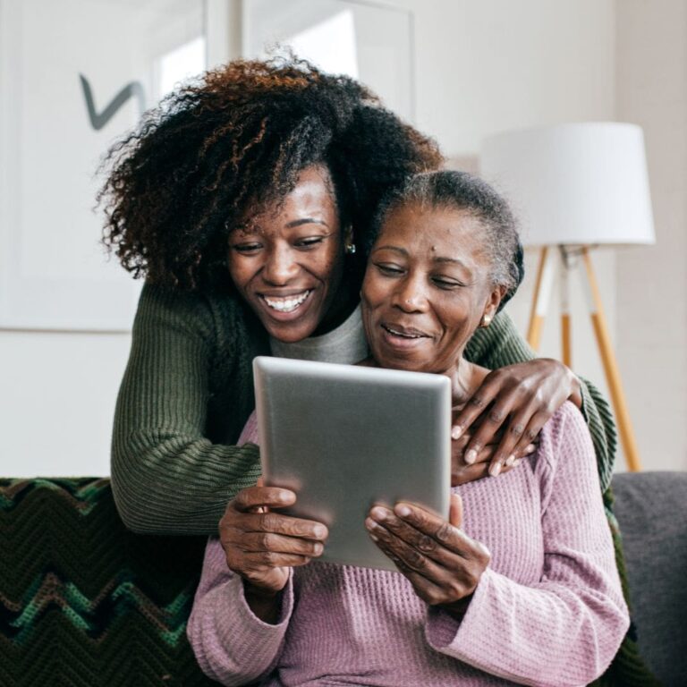 A smiling mother and daughter sitting together on a couch, looking at a tablet.
