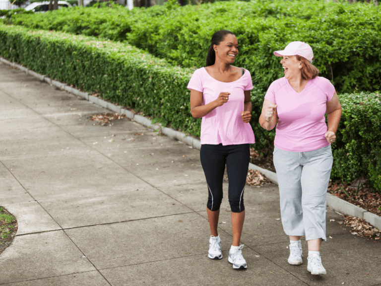 Two women wearing pink shirts walking and smiling on a paved path surrounded by greenery.