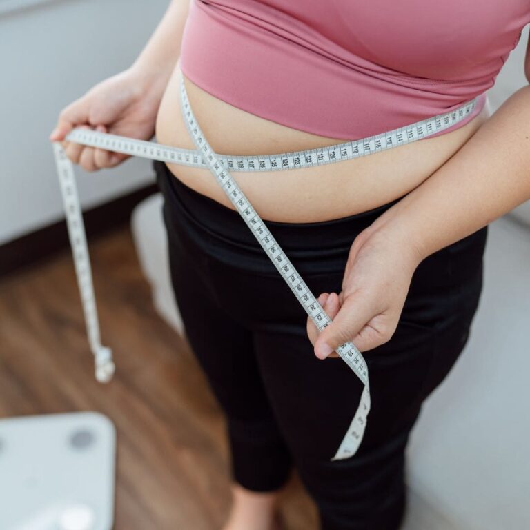 A woman in a pink top and black leggings measuring her waist with a tape measure.
